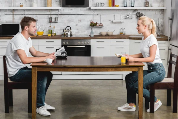 Vista lateral do jovem casal lendo e olhando um para o outro enquanto tomando café da manhã juntos — Fotografia de Stock
