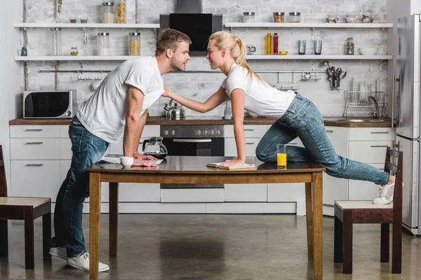 Junges leidenschaftliches Paar schaut einander an, junge Frau hält T-Shirt ihres hübschen Freundes in der Küche — Stockfoto