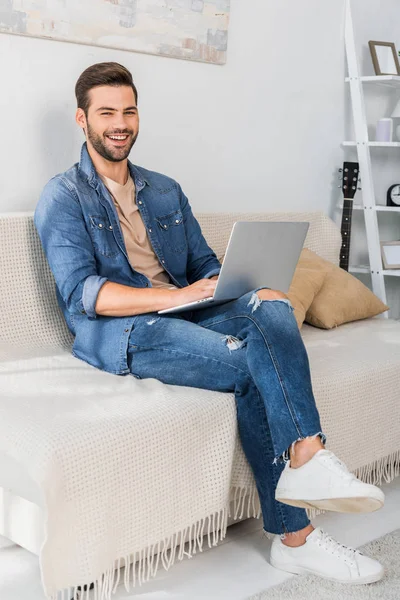 Сміється красивий молодий чоловічий фрілансер, використовуючи ноутбук на дивані вдома — стокове фото