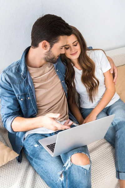 Vista de ángulo alto del hombre joven señalando con el dedo en la pantalla del ordenador portátil a la novia en el sofá en casa - foto de stock