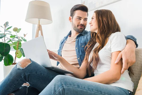 Bella giovane donna che punta sullo schermo del computer portatile al fidanzato sul divano a casa — Foto stock