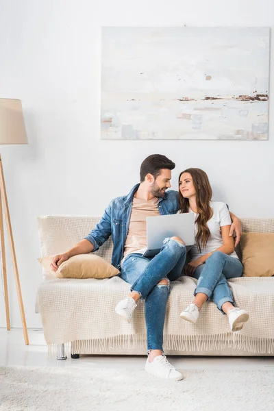 Joven feliz pareja con portátil sentado en el sofá y mirándose en casa - foto de stock