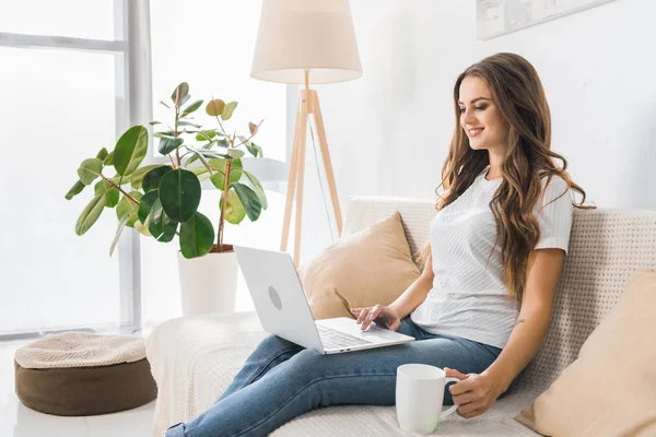 Mujer freelancer feliz con taza de café trabajando con el ordenador portátil en el sofá en casa - foto de stock