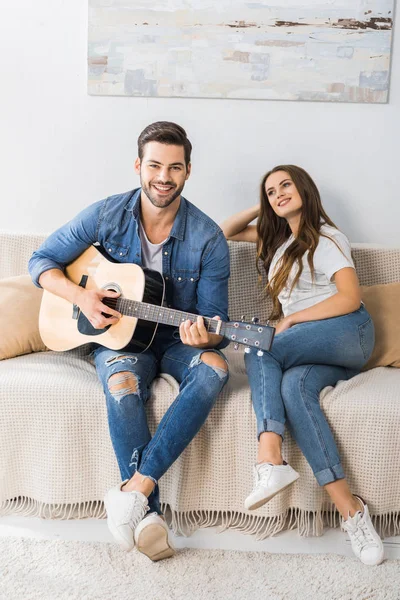 Lächelnder junger Mann spielt auf der Akustikgitarre, während seine Freundin in der Nähe auf der Couch zu Hause sitzt — Stockfoto