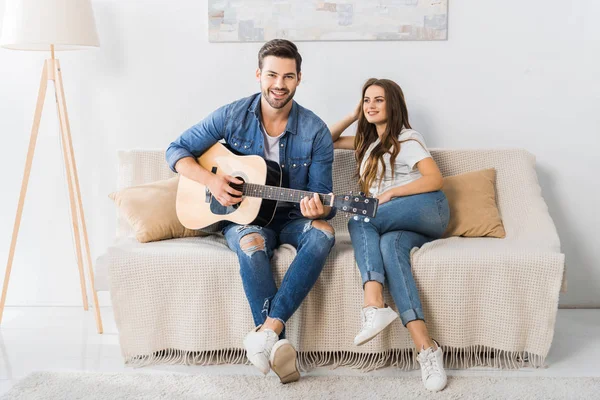 Молодий чоловік грає на акустичній гітарі, а його дівчина сидить поруч на дивані вдома — стокове фото