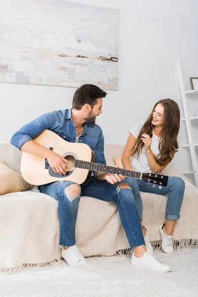 Pareja sonriente sentada en sofá con guitarra acústica en casa - foto de stock