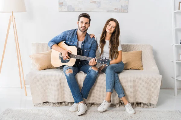 Glückliches junges Paar sitzt mit Akustikgitarre auf der Couch und schaut zu Hause in die Kamera — Stockfoto