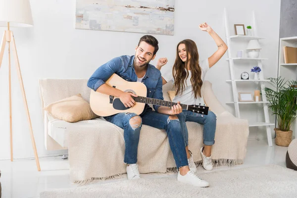 Sorrindo homem tocando na guitarra acústica, enquanto sua namorada dançando perto no sofá em casa — Fotografia de Stock