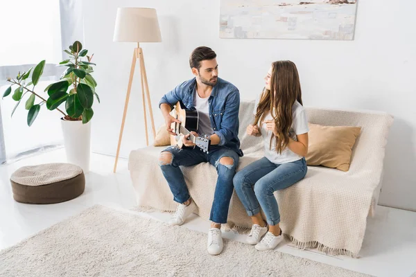 Joven hombre guapo tocando en la guitarra acústica mientras su novia bailando cerca en el sofá en casa - foto de stock