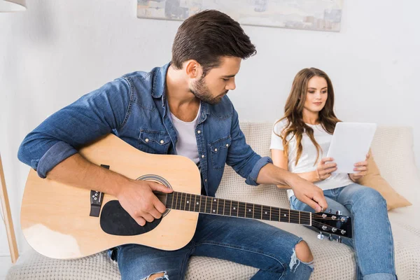 Handsome man tuning guitar while his girlfriend sitting near with digital tablet on sofa at home — Stock Photo