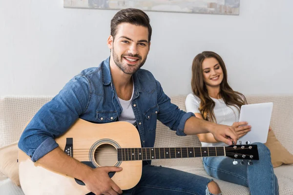 Smiling man tuning guitar and looking at camera while his girlfriend sitting near with digital tablet on sofa at home — Stock Photo