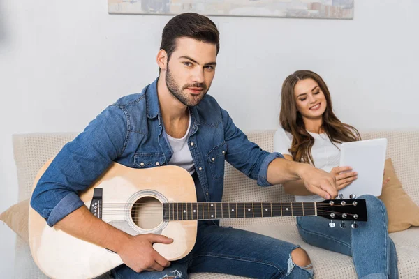 Homem confiante afinação guitarra e olhando para a câmera, enquanto sua namorada sentado perto com tablet digital no sofá em casa — Fotografia de Stock