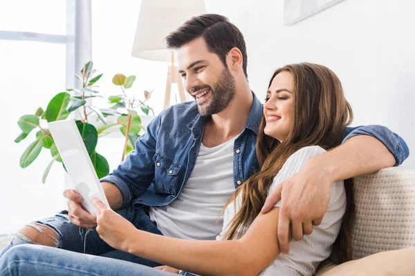 Riendo pareja joven sentada en el sofá con la tableta digital en casa - foto de stock