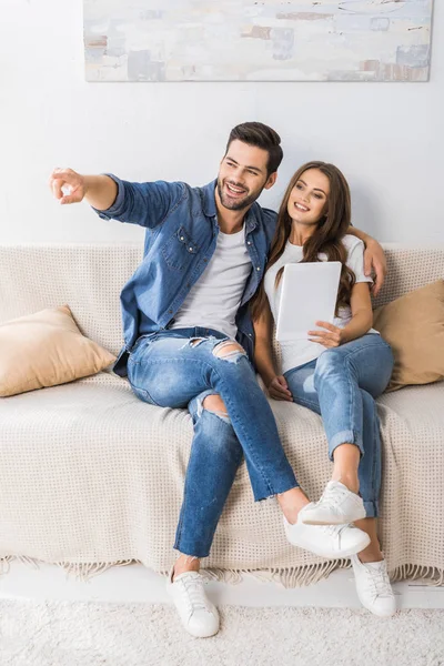 Smiling young man pointing by finger to girlfriend with digital tablet on couch at home — Stock Photo