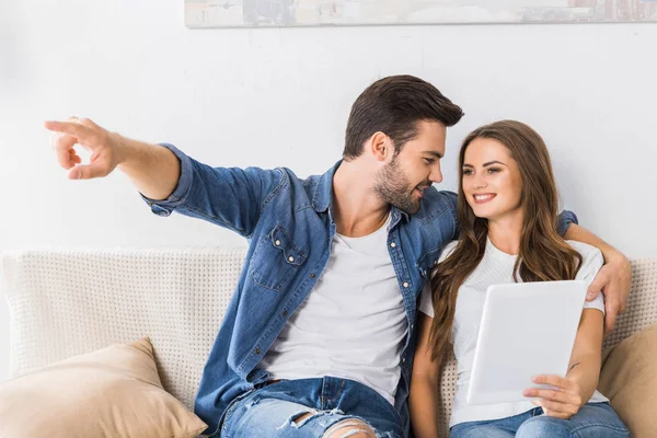 Jeune homme regardant petite amie avec tablette numérique et pointant du doigt vers elle sur le canapé à la maison — Photo de stock