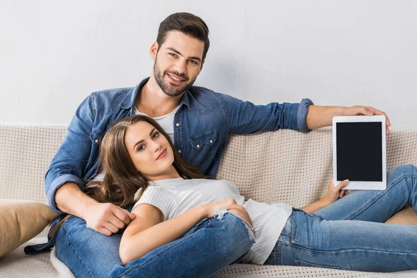 Lächelnde Frau zeigt digitales Tablet mit leerem Bildschirm, während ihr Freund in der Nähe auf der Couch zu Hause sitzt — Stockfoto