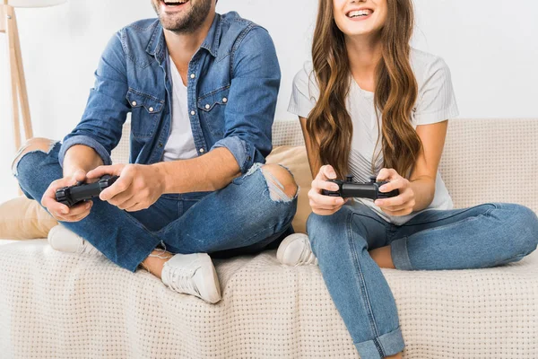 Visão parcial do casal jogando videogame por joysticks no sofá em casa — Fotografia de Stock