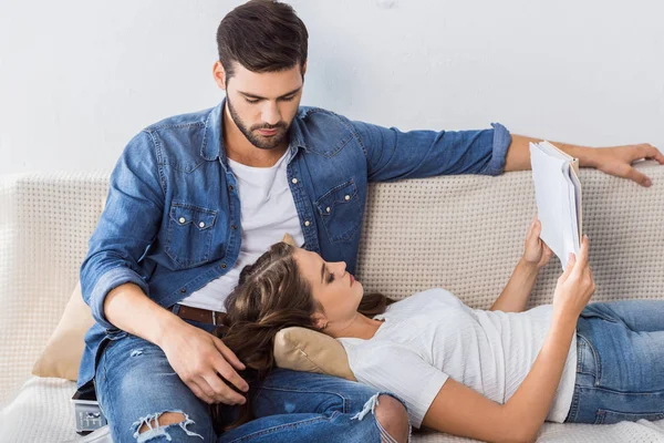 Young man touching girlfriend hair while she reading book on sofa at home — Stock Photo