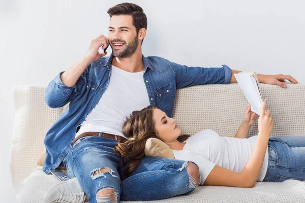 Homem feliz falando no smartphone enquanto sua namorada lendo livro no sofá em casa — Fotografia de Stock