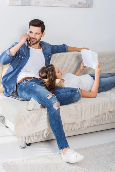Jovem homem bonito falando no smartphone enquanto sua namorada lendo livro no sofá em casa — Fotografia de Stock