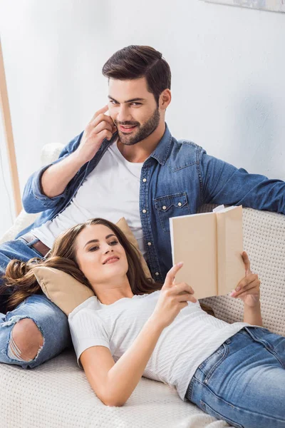 Smiling man talking on smartphone while his girlfriend reading book on sofa at home — Stock Photo