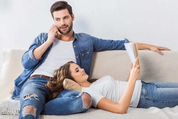 Joven guapo hablando en el teléfono inteligente, mientras que su novia leer libro en el sofá en casa - foto de stock
