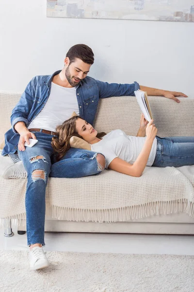 Lächelnder Mann mit Smartphone schaut Freundin an, während sie zu Hause auf der Couch Buch liest — Stockfoto