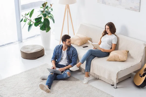 Vista de ángulo alto del hombre con el ordenador portátil y sentado en el suelo, mientras que su novia leer el libro en el sofá en casa - foto de stock