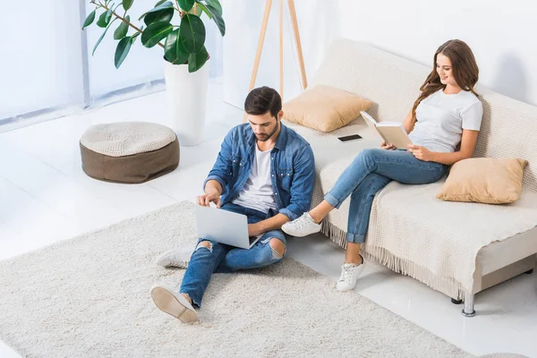 Vista de ángulo alto del hombre usando el ordenador portátil y sentado en el suelo, mientras que su novia leer libro en el sofá en casa - foto de stock