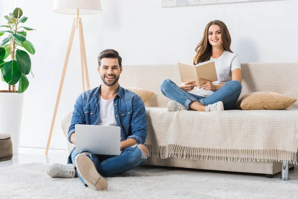 Sonriente hombre con portátil sentado en el suelo mientras su novia leer libro en el sofá en casa - foto de stock