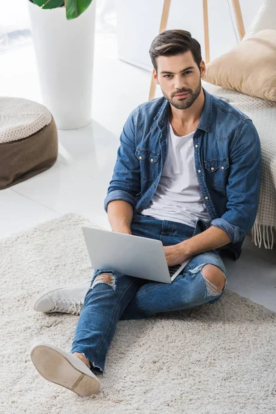 Vista de ángulo alto de guapo freelancer masculino trabajando con el ordenador portátil y mirando a la cámara en casa - foto de stock