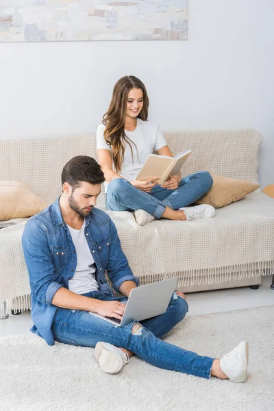 Hombre seguro de sí mismo con el ordenador portátil sentado en el suelo, mientras que su novia leer libro en el sofá en casa - foto de stock