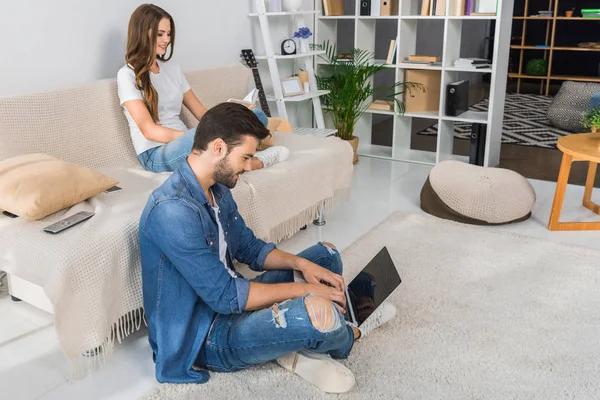 Vista lateral del hombre sonriente usando el ordenador portátil y sentado en el suelo, mientras que su novia leer libro en el sofá en casa - foto de stock