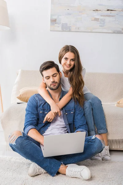 Smiling woman embracing boyfriend while he using laptop and sitting on floor at home — Stock Photo