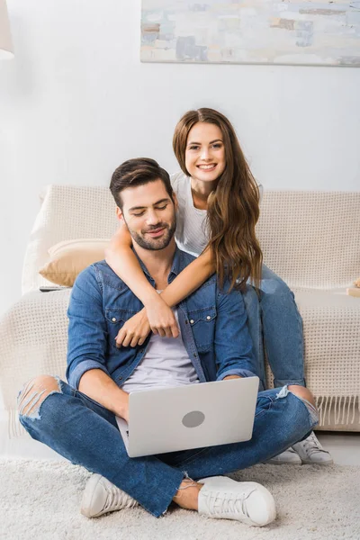 Atraente sorrindo mulher abraçando namorado enquanto ele usa laptop sentado no chão em casa — Fotografia de Stock