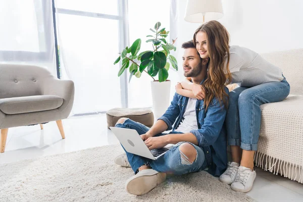 Happy young woman embracing boyfriend while he using laptop sitting on floor at home — Stock Photo