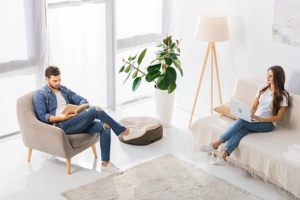 High angle view of woman using laptop on sofa while her boyfriend reading book on armchair at home — Stock Photo