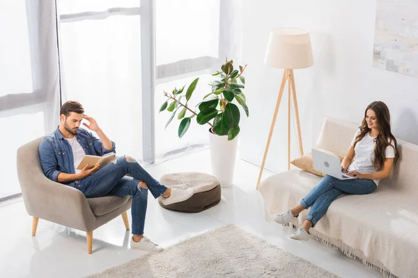 Aus der Vogelperspektive: Mann liest Buch auf Sessel, während seine Freundin Laptop auf Sofa zu Hause benutzt — Stockfoto