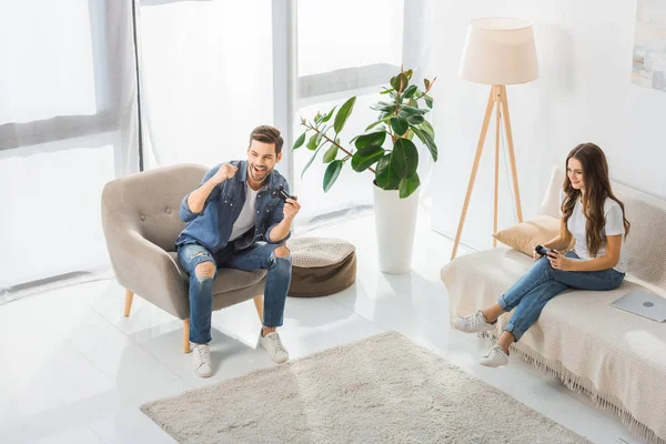 High angle view of happy man with joystick gesturing by hands and celebrating victory while his girlfriend sitting near on couch at home — Stock Photo