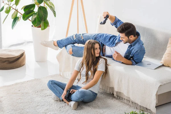 Young man threatening by joystick to smiling girlfriend sitting on floor at home — Stock Photo