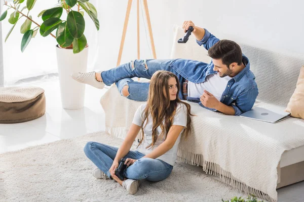 Joven amenazante por joystick a la novia sonriente sentada en el suelo en casa - foto de stock