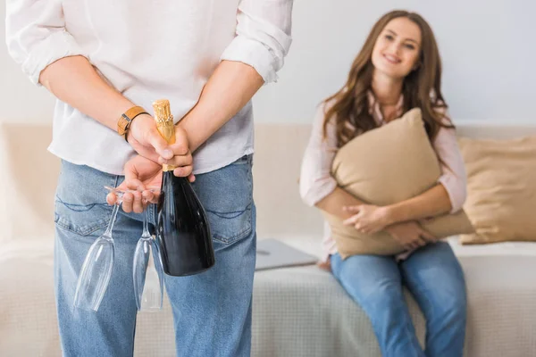 Partial view of man hiding bottle of champagne with glasses behind his back while his girlfriend sitting on couch at home — Stock Photo