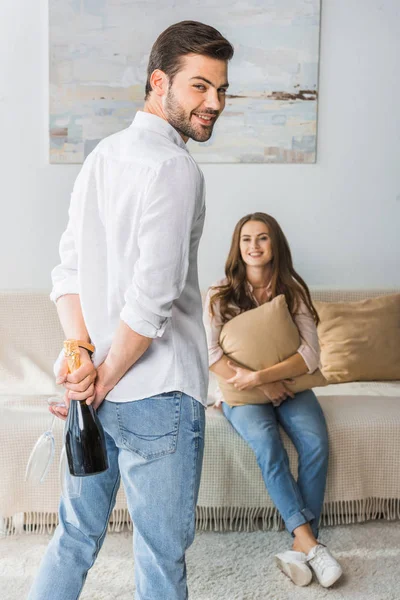 Smiling young man hiding bottle of champagne with glasses behind his back and looking at camera while his girlfriend sitting on couch at home — Stock Photo