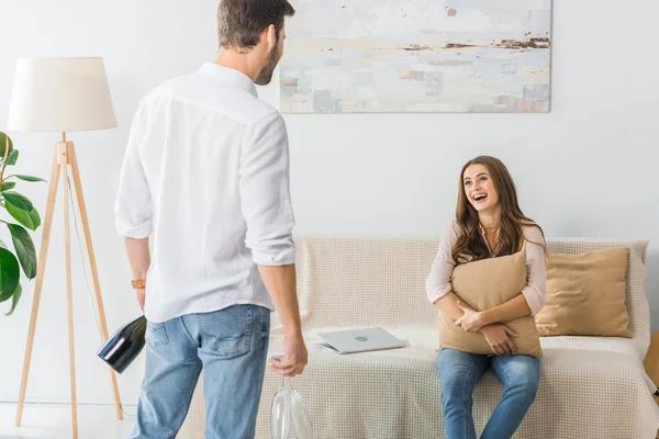 Vista posteriore dell'uomo in piedi con occhiali e bottiglia di champagne mentre la sua ragazza tiene il cuscino sul divano a casa — Foto stock