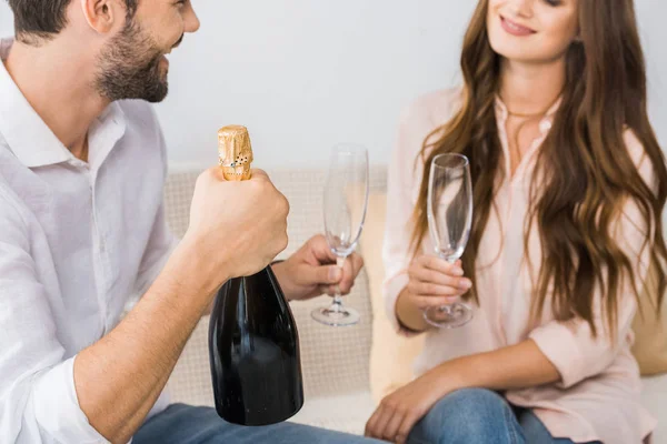 Vista parcial de la pareja sonriente celebrando con botella de champán y copas en el sofá en casa - foto de stock