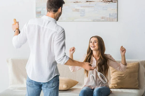 Vista trasera del hombre de pie con copas y botella de champán mientras su novia feliz gesticulando con las manos en el sofá en casa - foto de stock