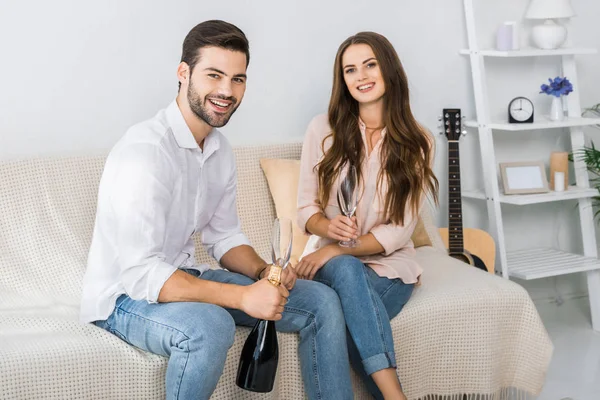 Heureux jeune couple célébrant avec bouteille de champagne et verres sur le canapé à la maison — Photo de stock