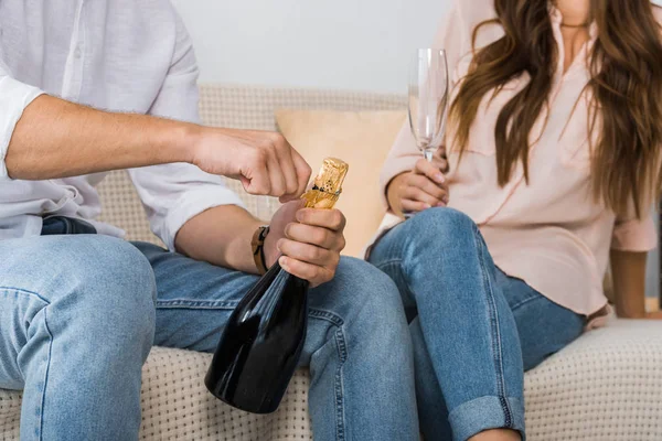 Imagem cortada de homem abrindo garrafa de champanhe, enquanto sua namorada sentada perto com vidro no sofá em casa — Fotografia de Stock