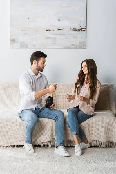 Hombre guapo abriendo botella de champán mientras su novia sentada cerca con vasos en el sofá en casa - foto de stock