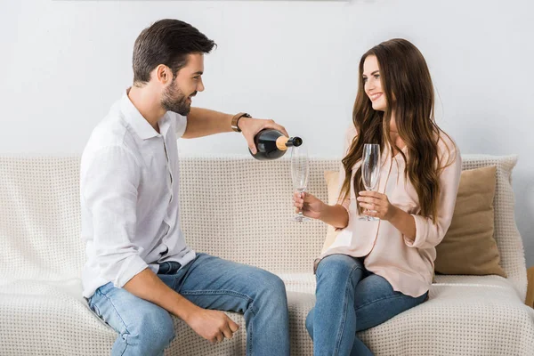 Vista lateral del hombre sonriente vertiendo champán en la copa mientras está sentado en el sofá con su novia en casa - foto de stock